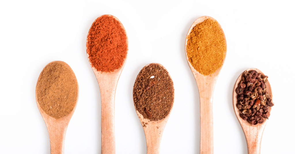 A row of wooden spoons with spices on a white background: cinnamon, paprika, curry, mixed spices, and Szechuan peppercorns.